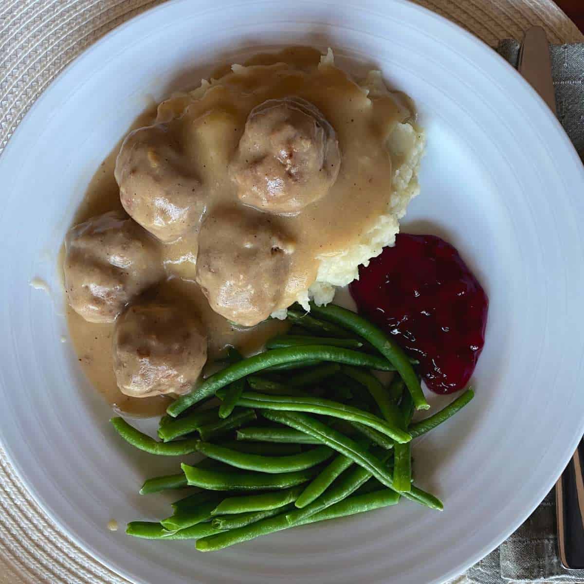 An overhead photo of a plate with gluten free Swedish meatballs on top of mashed potatoes with sides of green beans and lingonberry jam.