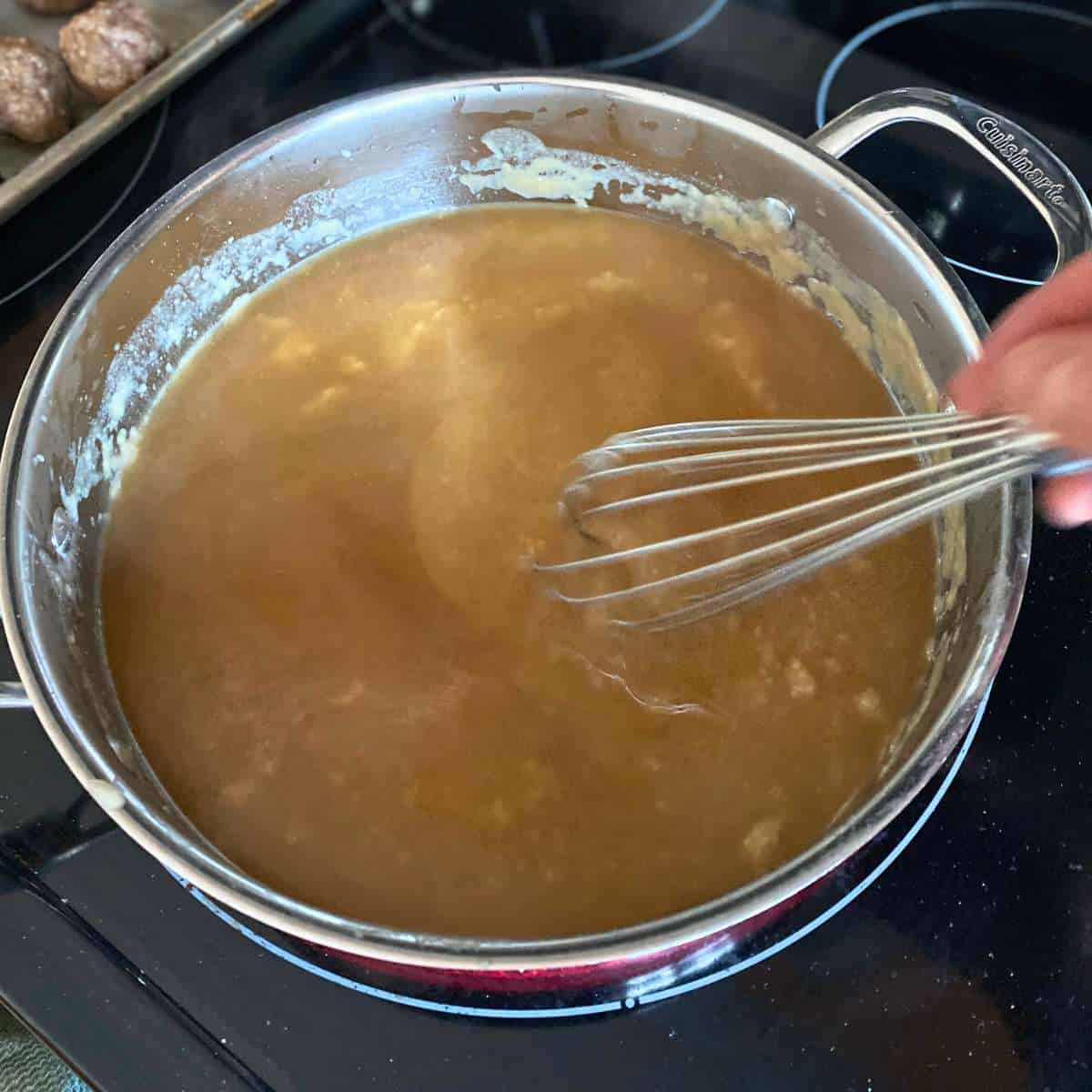 A large skillet with beef broth and roux being whisked together.